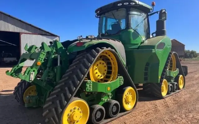 A John Deere 9570 RX quad track tractor with a front implement, parked on red soil beside a farm shed