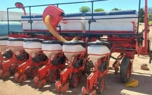 A red Kuhn six row planter with white seed hoppers, standing in a farmyard